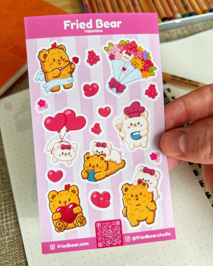 Hand holding the Fried Bear Valentine sticker sheet with hearts, balloons and a bouquet over a notebook